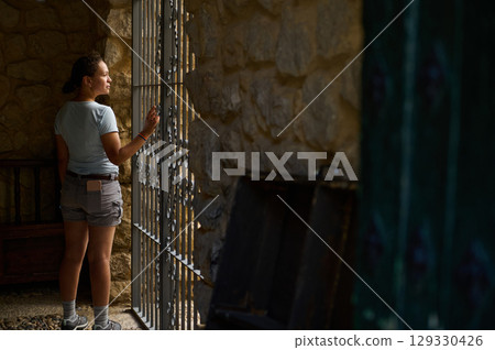 A Woman Admiring the View Through a Wrought Iron Window in a Rustic Stone Setting 129330426