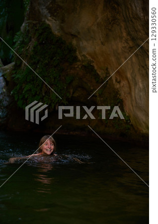 Cheerful Woman Swimming in Crystal Clear Natural Water Surrounded by Rock Formations and Green Foliage 129330560