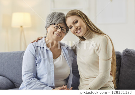 Smiling elderly mother and adult daughter sitting on sofa, hugging and looking at camera with love. 129331183