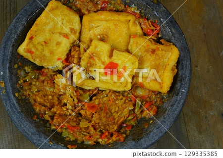 Tahu tempe penyet or Indonesian fried tofu and tempeh served on a dark stone mortar with a vibrant red chili paste known as sambal. 129335385