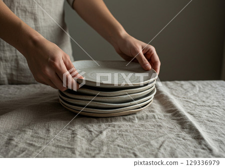 Womans Hands Stacking Ceramic Plates on Linen 129336979