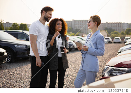 A couple happily receiving car keys from a salesperson, symbolizing a new car purchase at a dealership, captured outdoors. 129337348