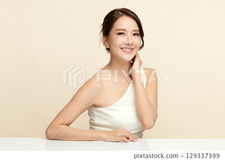 A beautiful young Asian woman with clean, fresh skin against a beige background. Skincare, facial treatment, cosmetology, beauty, and spa. An elegant portrait of an Asian woman. 129337399