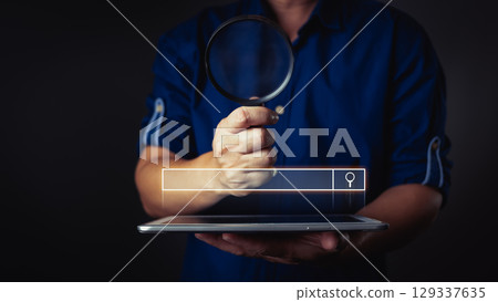 A person holds a magnifying glass over a tablet, symbolizing search, inquiry, and digital exploration in a dark background. 129337635