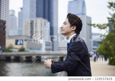 A Japanese businessman in a suit gazing into the distance with a building in the background A Japanese businessman in a suit gazing into the distance with a building in the background 129337829