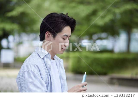 A young Japanese man rests on a park bench, holding an e-cigarette. A young Japanese man rests on a park bench, holding an e-cigarette. 129338217