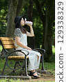 A young woman sitting on a bench in the shade of a tree and drinking water from her own bottle 129338329