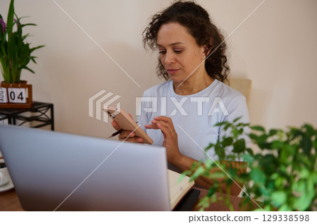 Woman Working Remotely Using Smartphone and Laptop in Cozy Home Environment Woman Working Remotely Using Smartphone and Laptop in Cozy Home Environment 129338598