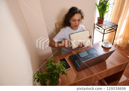 Remote Work Setup Featuring Woman with Notebook and Laptop in Cozy Home Office Remote Work Setup Featuring Woman with Notebook and Laptop in Cozy Home Office 129338606