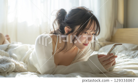 A woman lying face down on the bed and looking at her smartphone 129338867