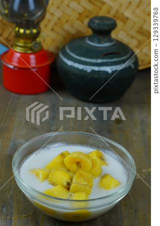 A glass bowl of kolak pisang or Indonesian banana compote in coconut milk, with a red oil lamp and a traditional clay pot in the background. A glass bowl of kolak pisang or Indonesian banana compote in coconut milk, with a red oil lamp and a traditional clay pot in the background. 129339768
