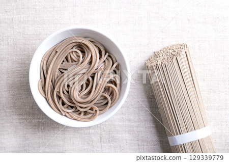 Soba noodles, boiled and chilled, in white bowl on linen fabric. Thin Japanese noodles, made primarily from buckwheat flour, with wheat flour mixed in. With tied bundle of dried noodles on the right. Soba noodles, boiled and chilled, in white bowl on linen fabric. Thin Japanese noodles, made primarily from buckwheat flour, with wheat flour mixed in. With tied bundle of dried noodles on the right. 129339779