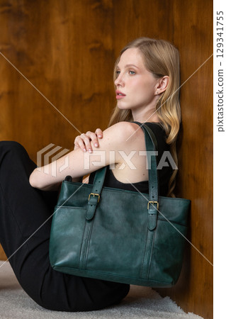A fashion model with long wavy blonde hair is captured in a full-length, grounded pose, resting against a textured wood-paneled wall while showcasing a dark teal leather tote bag 129341755