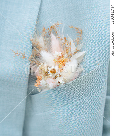 A delicate arrangement of dried flowers and grasses tucked neatly into the pocket of a light blue suit, creating a charming accent perfect for formal occasions and weddings. 129341784