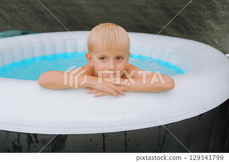 Young boy leaning on the edge of an inflatable pool, looking relaxed and thoughtful. The outdoor scene captures a peaceful moment, emphasizing leisurely enjoyment in a casual setting. Young boy leaning on the edge of an inflatable pool, looking relaxed and thoughtful. The outdoor scene captures a peaceful moment, emphasizing leisurely enjoyment in a casual setting. 129341799