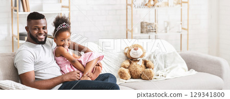 African American father is sitting on a comfortable couch with his daughter, who is playfully leaning against him. They are enjoying a tender moment together, copy space African American father is sitting on a comfortable couch with his daughter, who is playfully leaning against him. They are enjoying a tender moment together, copy space 129341800