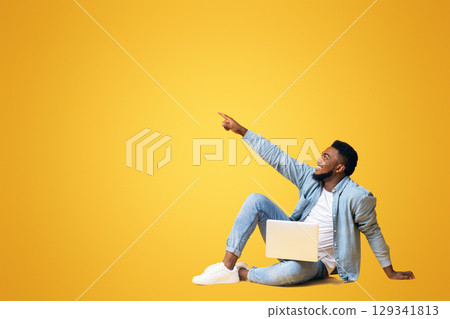 Look there. African american guy sitting on floor with laptop and pointing aside at copy space on yellow background 129341813