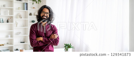 A cheerful Indian man with long curly hair and glasses stands with arms crossed, exuding confidence and warmth. The bright room is decorated with natural light, copy space A cheerful Indian man with long curly hair and glasses stands with arms crossed, exuding confidence and warmth. The bright room is decorated with natural light, copy space 129341873