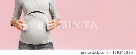 Music and pregnancy. Expectant woman holding headphones near belly, pink studio background, crop 129341889