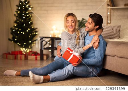 Handsome guy surprising girlfriend with Christmas present, resting together at home, free space 129341899
