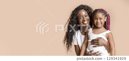 Mommy's girl. Happy african american mother embracing her adorable little daughter, yellow studio background 129341929