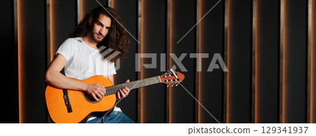 A young musician with long hair passionately strums an acoustic guitar in a modern studio setting. The warm light highlights his focused expression as he engages with his music, copy space A young musician with long hair passionately strums an acoustic guitar in a modern studio setting. The warm light highlights his focused expression as he engages with his music, copy space 129341937