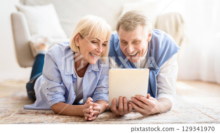 Excited mature couple browsing social media on tablet, lying on carpet at home 129341970