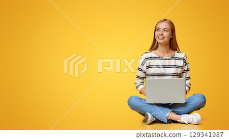 Dreamy teenage girl sitting with laptop on floor and looking aside at copy space on orange background 129341987