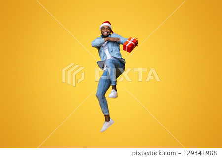 Christmas delivery concept. Positive african american guy jumping with Xmas present, hurry to deliver gift box in time. yellow background 129341988