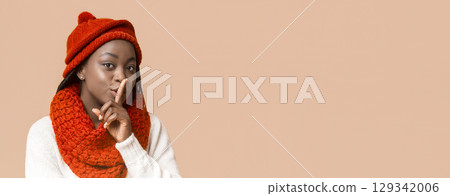 A young black woman softly gestures for silence, wearing a cozy red hat and matching scarf. She holds her finger to her lips, conveying a sense of calm and secrecy, copy space 129342006