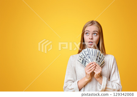 Thoughtful Girl Holding Bunch Of Dollars And Looking Upwards On Orange Studio Background 129342008