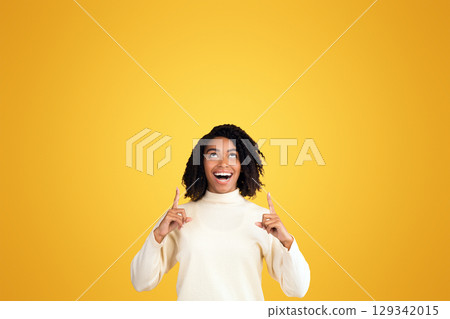Place For Ad. Portrait of happy black girl pointing fingers up at copyspace isolated over yellow background, vertical image 129342015