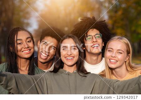 Smiling teen international friends taking selfie while walking in autumn park, happy memories concept 129342017