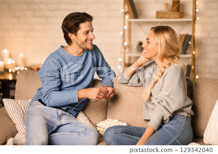 Happy millennial couple talking and eating popcorn in cozy living room, free space 129342040