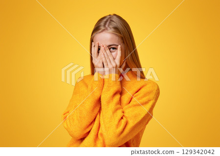 Shy teenage girl peeking through fingers, covering face with both hands over orange studio background with copy space Shy teenage girl peeking through fingers, covering face with both hands over orange studio background with copy space 129342042