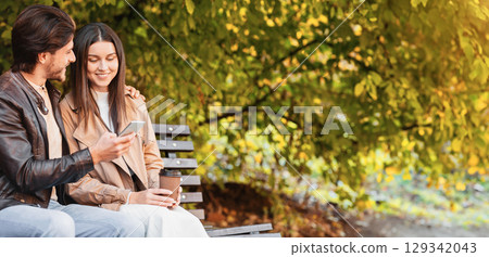 Man and woman sitting on bench and watching photos on mobile phone, spending time together in park, copy space 129342043