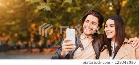 Young happy lovers enjoying time together, taking selfie while walking by golden forest, using mobile phone, close up Young happy lovers enjoying time together, taking selfie while walking by golden forest, using mobile phone, close up 129342080