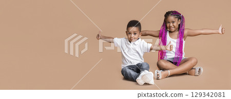 We like it. Happy little african american kids, sister and brother, showing thumbs up gesture, sitting with hands spread, yellow studio background 129342081