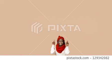 A cheerful young African American woman with a big smile is wearing a cozy red hat and matching scarf. She is enthusiastically pointing upward at copy space 129342082