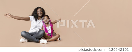 Like mother like daughter. Cute little afro girl sitting on floor with mom and repeating her gestures, yellow background with free space 129342101
