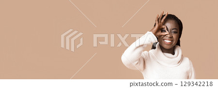 A joyful African American woman is playfully posing by forming a circle with her hand, smiling brightly. She is dressed in a cozy sweater, and the background features a soft, neutral tone, copy space A joyful African American woman is playfully posing by forming a circle with her hand, smiling brightly. She is dressed in a cozy sweater, and the background features a soft, neutral tone, copy space 129342218