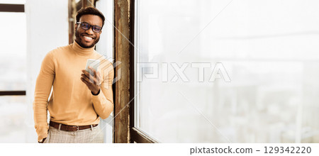 Modern Technologies At Work. Happy african guy using phone in front of window. Panorama, empty space Modern Technologies At Work. Happy african guy using phone in front of window. Panorama, empty space 129342220