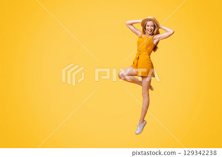 Summer Fashion. Joyful Teen Girl In Jumpsuit And Straw Hat Jumping On Yellow Studio Background With Copy Space 129342238