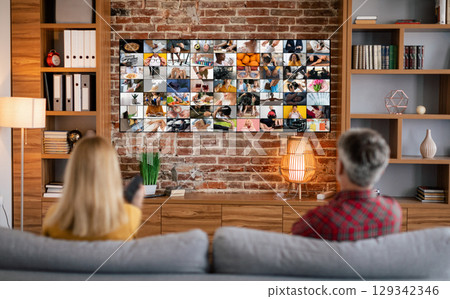 A couple relaxes on a couch in their living room, facing a large television screen displaying a grid of various images. The room features wooden shelves filled with books and decor, a brick wall A couple relaxes on a couch in their living room, facing a large television screen displaying a grid of various images. The room features wooden shelves filled with books and decor, a brick wall 129342346