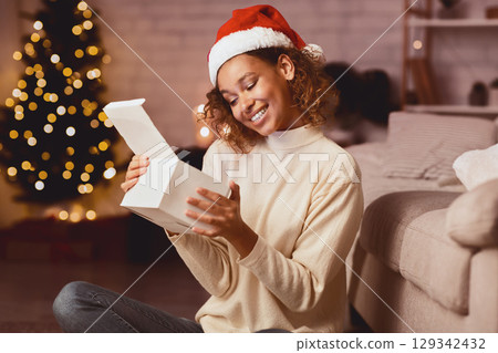 Christmas. African-american woman opening present in front of Christmas tree at home 129342432