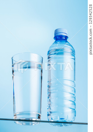 Glass of fresh water and bottle on a glass table against a vibrant blue background 129342528
