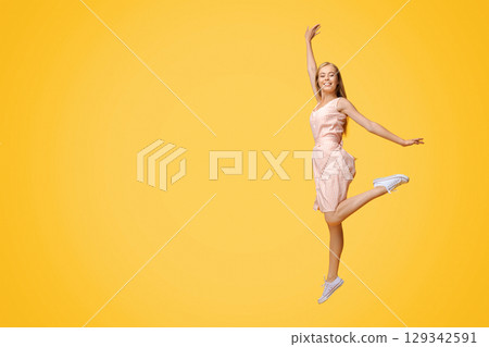 Full Length Portrait Of Joyful Teen Girl In Summer Cocktail Dress Jumping High In The Air Over Yellow Background In Studio, Copy Space 129342591