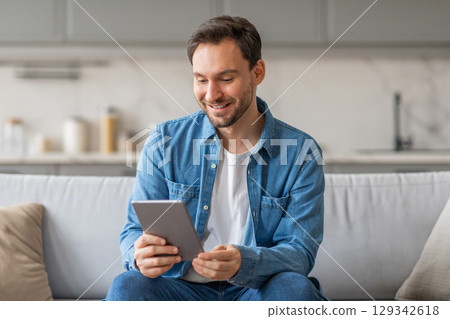 A man in a blue shirt and jeans sits on a light gray couch in a living room. He is looking down at a tablet, smiling, and appears to be browsing the internet or using an app. 129342618