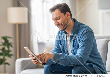 A man is sitting on a sofa in a living room, smiling and using a tablet. The sofa is a light grey color and the walls are a soft white. He is wearing a blue denim shirt and blue jeans. 129342629