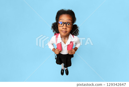 Shocked African School Girl Looking At Camera Standing With Backpack Over Yellow Studio Background. High-Angle, Vertical Shocked African School Girl Looking At Camera Standing With Backpack Over Yellow Studio Background. High-Angle, Vertical 129342647